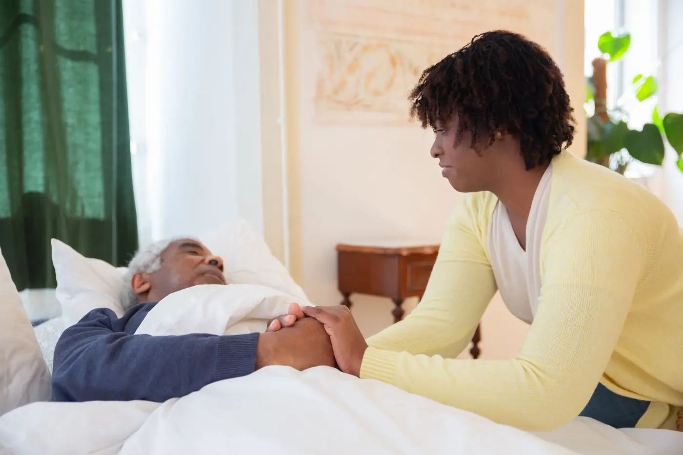 A carer gently holding an older man&rsquo;s hand as he rests in bed